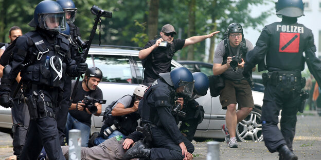 Polizisten drücke einen Menschen zu Boden daneben steht ein gestikulierender Journalist Polizisten drücke einen Menschen zu Boden daneben steht ein gestikulierender Journalist