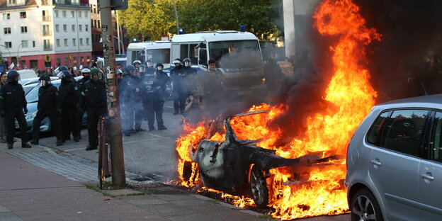 Ein brennendes Auto und Polizisten im Hintergrund Ein brennendes Auto und Polizisten im Hintergrund