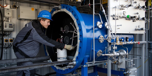 Ein Mann mit Helm beuht sich über ein technisches Gerät, das aus einer blauen Röhre ragt Ein Mann mit Helm beuht sich über ein technisches Gerät, das aus einer blauen Röhre ragt