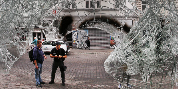 Zwei Männer sind im Hamburger Schanzenviertel auf der Straße durch eine zerbrochene Glasscheibe hindurch zu sehen Zwei Männer sind im Hamburger Schanzenviertel auf der Straße durch eine zerbrochene Glasscheibe hindurch zu sehen