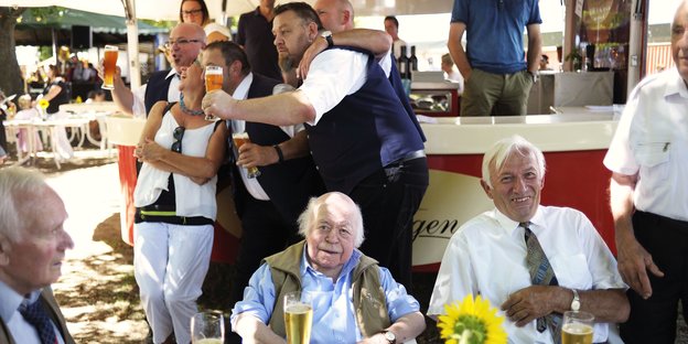 Drei ältere Männer sitzen an einem Tisch und trinken Bier, dahinter posieren Menschen vor einem Getränkestand für ein Foto
