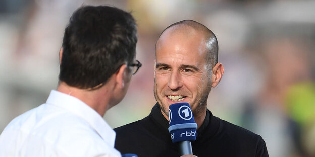 Ein Mann, Mehmet Scholl, hält ein Mikrofon mit dem ARD-Logo, neben ihm ein weiterer Mann Ein Mann, Mehmet Scholl, hält ein Mikrofon mit dem ARD-Logo, neben ihm ein weiterer Mann
