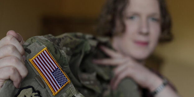 Eine Frau mit kurzen Haaren hält eine Militär-Jacke mit US-amerikanischer Flagge auf dem Arm hoch Eine Frau mit kurzen Haaren hält eine Militär-Jacke mit US-amerikanischer Flagge auf dem Arm hoch