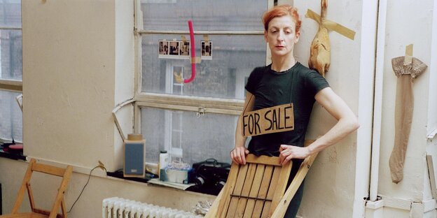 Eine Frau lehnt an einer Wand, um ihren Hals hängt ein Schild mit der Aufschrift "For Sale" Eine Frau lehnt an einer Wand, um ihren Hals hängt ein Schild mit der Aufschrift "For Sale"