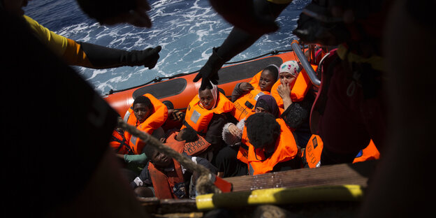 Menschen in einem Schlauchboot, davor als Silhouetten HelferInnen Menschen in einem Schlauchboot, davor als Silhouetten HelferInnen