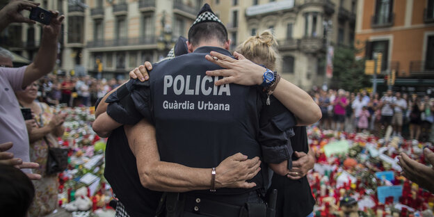 Ein Polizist ist von hinten zu sehen, er umarmt zwei Menschen, im Hintergrund sind Kerzen und Blumen aufgestellt Ein Polizist ist von hinten zu sehen, er umarmt zwei Menschen, im Hintergrund sind Kerzen und Blumen aufgestellt