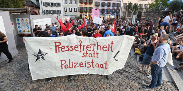 Menschen mit großem Transparent mit der Aufschrift "Pressefreiheit statt Polizeistaat" auf einem Platz in einem Ort Menschen mit großem Transparent mit der Aufschrift "Pressefreiheit statt Polizeistaat" auf einem Platz in einem Ort