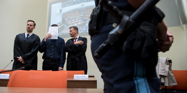 Ein Mann hält ein Papier vor sein Gesicht, neben ihm stehen zwei Männer, ein davon in einer Robe. Im Vordergrund steht ein Justizbeamter Ein Mann hält ein Papier vor sein Gesicht, neben ihm stehen zwei Männer, ein davon in einer Robe. Im Vordergrund steht ein Justizbeamter