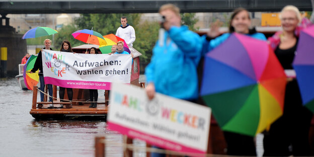 Menschen auf Flößen in einem Fluß, sie tragen bunte Regenschirme und ein Transparent mit der Aufschrift "Aufwachen: Wählen gehen" Menschen auf Flößen in einem Fluß, sie tragen bunte Regenschirme und ein Transparent mit der Aufschrift "Aufwachen: Wählen gehen"
