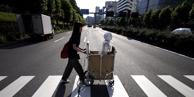 Eine Frau schiebt einen Roboter in einem Wagen über eine Straße Eine Frau schiebt einen Roboter in einem Wagen über eine Straße