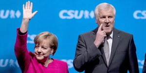 Merkel in Pink reckt die rechte Hand in die Luft, Seehofer berührt mit dem Zeigefinger seine Wange