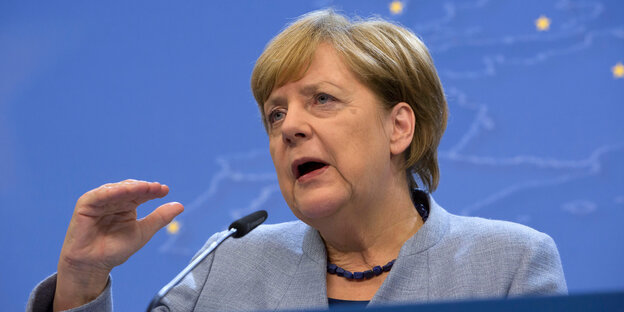 Angela Merkel spricht in ein Mikrofon und hebt die Hand Angela Merkel spricht in ein Mikrofon und hebt die Hand