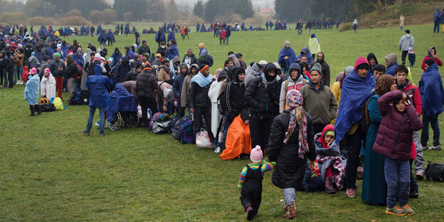Flüchtlinge in mehr oder weniger warmer Kleidung stehen auf einer kalt und verregnet aussehenden Wiese Flüchtlinge in mehr oder weniger warmer Kleidung stehen auf einer kalt und verregnet aussehenden Wiese