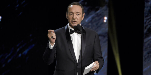 Kevin Spacey im Anzug, eine Hand erhoben, in der anderen ein paar Papiere