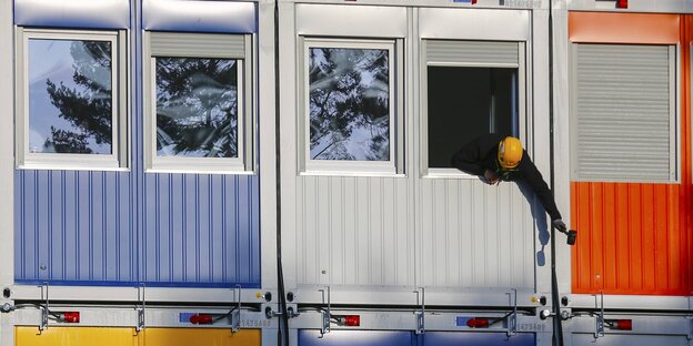 Mehrere Wohncontainer übereinandergestapelt. Aus einem Fenster beugt sich ein Mann mit Bauarbeiterhelm