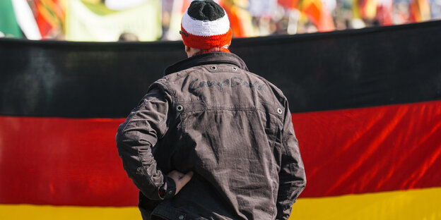 Ein Mann mit Strickmütze in den Nationalfarben des deutschen Reiches steht vor einer Deutschlandfahne Ein Mann mit Strickmütze in den Nationalfarben des deutschen Reiches steht vor einer Deutschlandfahne