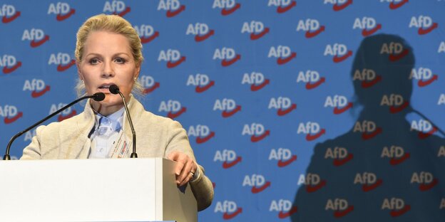 Frau mit blonden Haaren am Redepult, rechts ihr Schatten, dahinter blaue Tapete mit Aufschrift "AfD" Frau mit blonden Haaren am Redepult, rechts ihr Schatten, dahinter blaue Tapete mit Aufschrift "AfD"