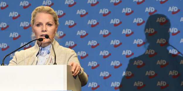 Frau mit blonden Haaren am Redepult, rechts ihr Schatten, dahinter blaue Tapete mit Aufschrift "AfD"