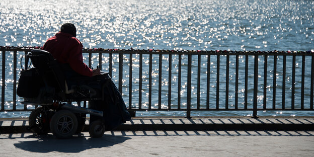 Eine Frau in einem Rollstuhl schaut von einer Promenade aufs Meer Eine Frau in einem Rollstuhl schaut von einer Promenade aufs Meer