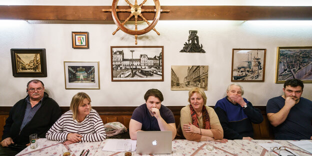 Menschen sitzen nebeneinander mit nachdenklichen Gesichtern an einem Kneipentisch, auf dem ein Laptop steht