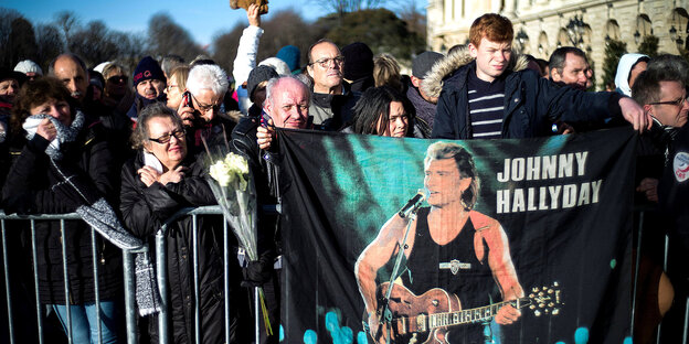 Mehrere Menschen an einem Sperrgitter, darüber hängt ein Transparent mit Johnny Hallyday Mehrere Menschen an einem Sperrgitter, darüber hängt ein Transparent mit Johnny Hallyday