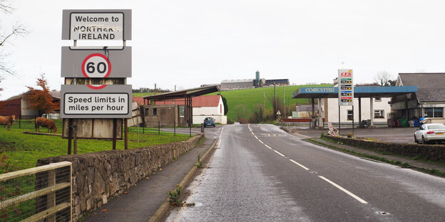 Eine Straße, die in ein Dorf an einem Hügel führt. Links ein Orts- und Geschwindigkeitsbegrenzungsschild Eine Straße, die in ein Dorf an einem Hügel führt. Links ein Orts- und Geschwindigkeitsbegrenzungsschild