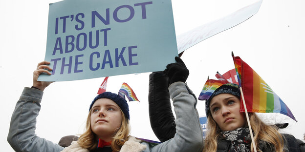 Zwei Demonstrantinnen, eine mit Regenbogenflagge, die andere hält ein Schild: "It's not about the cake" Zwei Demonstrantinnen, eine mit Regenbogenflagge, die andere hält ein Schild: "It's not about the cake"
