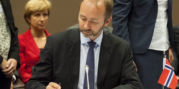 Trond Giske sitzt an einem Tisch mit einer kleinen norwegischen Flagge und unterschreibt Dokumente Trond Giske sitzt an einem Tisch mit einer kleinen norwegischen Flagge und unterschreibt Dokumente