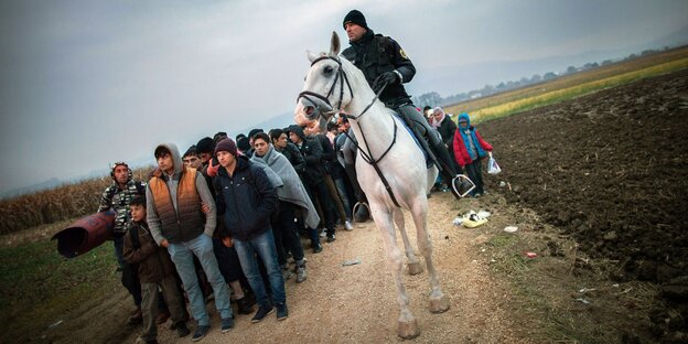 Flüchtlinge stehen auf einem Feld. Neben ihnen ein Polizist auf einem Pferd Flüchtlinge stehen auf einem Feld. Neben ihnen ein Polizist auf einem Pferd