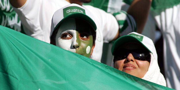 zwei weibliche Fans mit Basecaps und saudi-arabischer Fahne, eine mit grün-weiß angemaltem Gesicht zwei weibliche Fans mit Basecaps und saudi-arabischer Fahne, eine mit grün-weiß angemaltem Gesicht