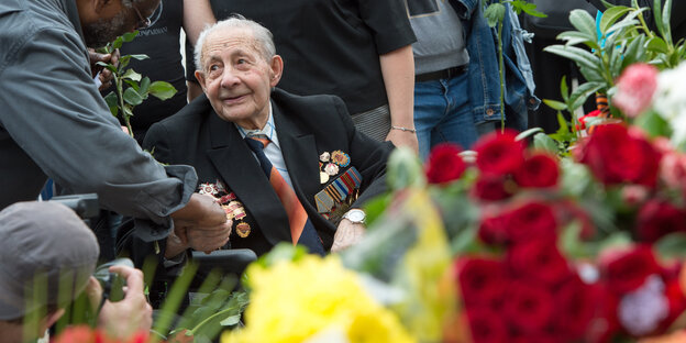 Ein alter Mann sitzt schräg hinter Rosen. Jemand schüttelt ihm die Hand Ein alter Mann sitzt schräg hinter Rosen. Jemand schüttelt ihm die Hand