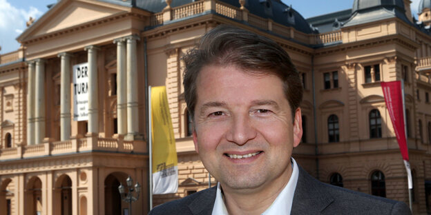 Ein mittelalter Mann mit Hemd und Sakko steht vor der Fassade des Mecklenburgischen Staatstheaters. Ein mittelalter Mann mit Hemd und Sakko steht vor der Fassade des Mecklenburgischen Staatstheaters.