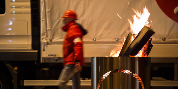 Ein Mann in roter Jacke und rotem Käppi geht hinter einer brennenden Feuertonne entlang Ein Mann in roter Jacke und rotem Käppi geht hinter einer brennenden Feuertonne entlang