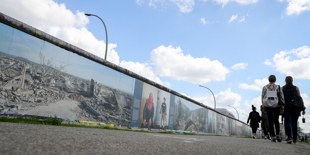 Leute laufen neben einem Stück Berliner Mauer lang Leute laufen neben einem Stück Berliner Mauer lang