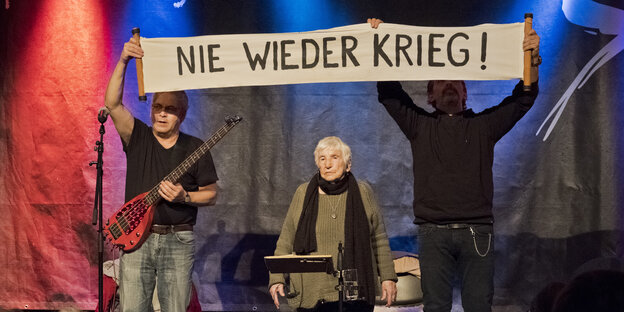 Zwei mittelalte Männer halten ein Plakat, auf dem steht: "Nie wieder Krieg!" Zwischen den Männern steht eine 93-Jährige Frau hinter einem Notenständer. Zwei mittelalte Männer halten ein Plakat, auf dem steht: "Nie wieder Krieg!" Zwischen den Männern steht eine 93-Jährige Frau hinter einem Notenständer.