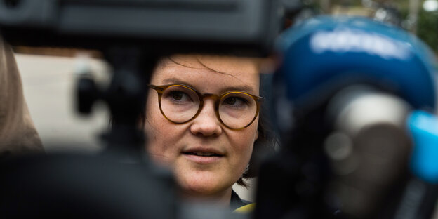 Eine junge Frau mit dunklen Haaren und dunkler Brille wird umringt von Mikrophonen. Eine junge Frau mit dunklen Haaren und dunkler Brille wird umringt von Mikrophonen.