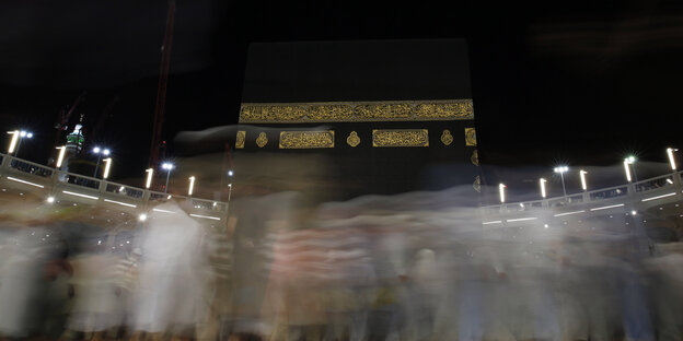 Im Vordergrund verschwommene Silhouetten vorbeilaufender Menschen, im Hintergrund die Kaaba vor dem schwarzen Nachthimmel. Im Vordergrund verschwommene Silhouetten vorbeilaufender Menschen, im Hintergrund die Kaaba vor dem schwarzen Nachthimmel.