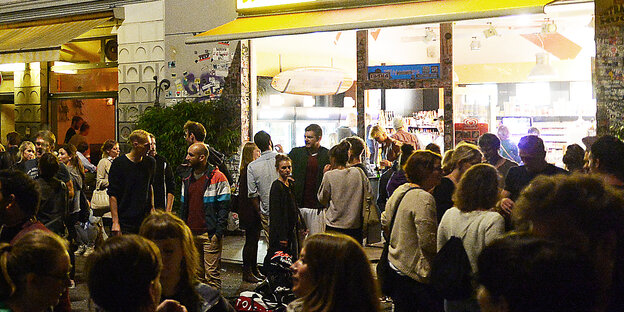 Junge Leute stehen in kleinen Gruppen vor einem Kiosk auf der Straße und reden miteinander.