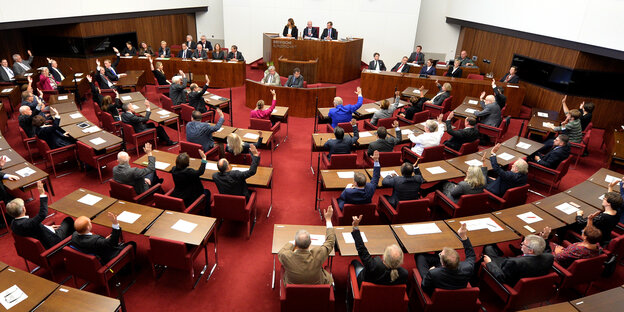Abgeordnete sitzen hinter braunen Tischen und heben die Hand zur Abstimmung. Abgeordnete sitzen hinter braunen Tischen und heben die Hand zur Abstimmung.