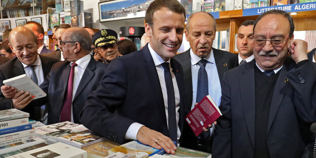 Macron steht lächelnd in einem Buchladen, um ihn herum viele ältere Männer Macron steht lächelnd in einem Buchladen, um ihn herum viele ältere Männer