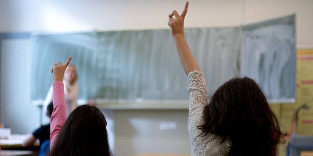 Zwei Schülerinnen strecken in der Klasse den Arm in die Höhe, einen Lehrerin steht an der Tafel Zwei Schülerinnen strecken in der Klasse den Arm in die Höhe, einen Lehrerin steht an der Tafel
