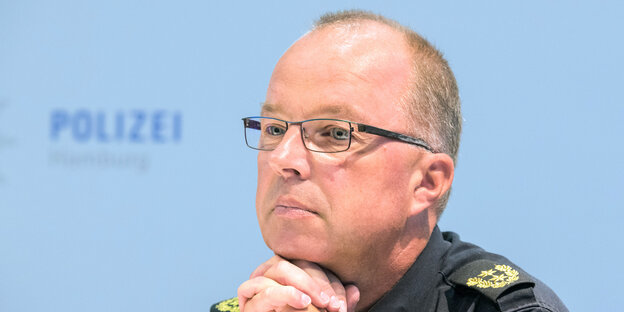 Ein Mann mit hoher Stirn, kurzen grau-blonden Haaren und einer dunklen Brille sitzt vor einer blauen Wand, auf der "Polizei" steht. Ein Mann mit hoher Stirn, kurzen grau-blonden Haaren und einer dunklen Brille sitzt vor einer blauen Wand, auf der "Polizei" steht.