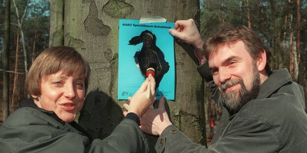Jochen Flasbarth steht mit Angela Merkel vor einem Baum und hält ein Plakat fest, während sie darauf herumpinselt Jochen Flasbarth steht mit Angela Merkel vor einem Baum und hält ein Plakat fest, während sie darauf herumpinselt
