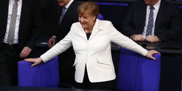Eine Frau in weißem Blazer breitet ihre Arme aus und hält sich so an zwei blauen Stühlen fest Eine Frau in weißem Blazer breitet ihre Arme aus und hält sich so an zwei blauen Stühlen fest