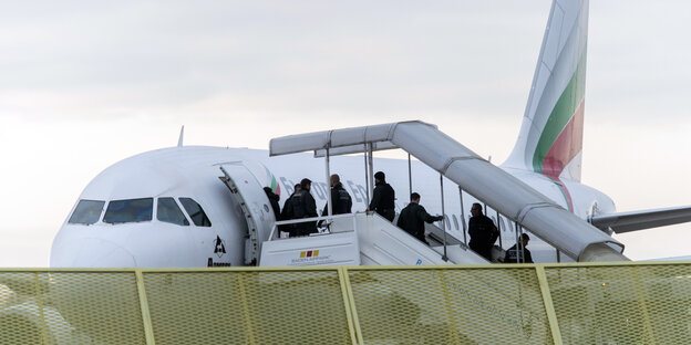 Über eine Treppe besteigen mehrere Menschen ein Flugzeug. Polizisten überwachen sie dabei.