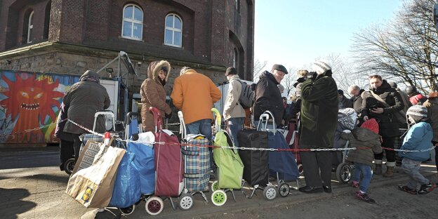 Menschen warten mit Einkaufsbeuteln vor einem Gebäude auf die Öffnung der Tafel Menschen warten mit Einkaufsbeuteln vor einem Gebäude auf die Öffnung der Tafel