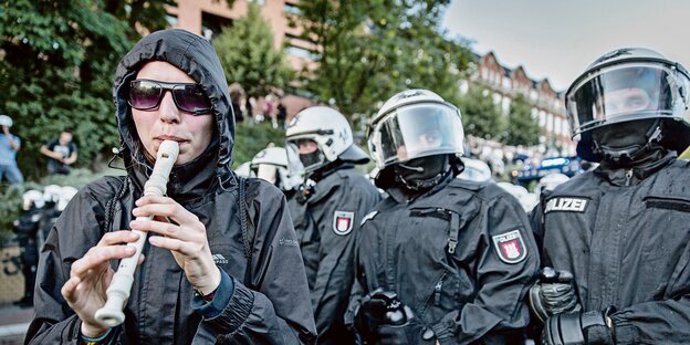 eine Demonstrantin spielt auf einer Blockflöte - dahinter behelmte Polizisten mit geschlossenem Visier eine Demonstrantin spielt auf einer Blockflöte - dahinter behelmte Polizisten mit geschlossenem Visier