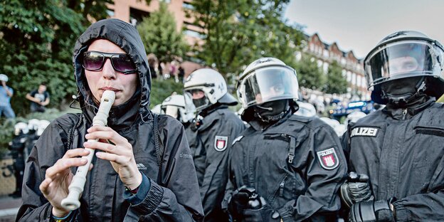 eine Demonstrantin spielt auf einer Blockflöte - dahinter behelmte Polizisten mit geschlossenem Visier