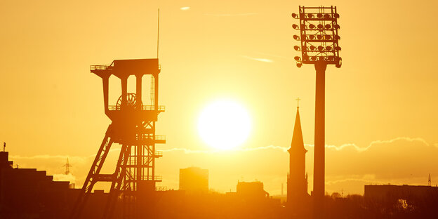 Die Sonne geht über dem Förderturm einer Zeche und einem Flutlichtmast eines Stadion auf Die Sonne geht über dem Förderturm einer Zeche und einem Flutlichtmast eines Stadion auf