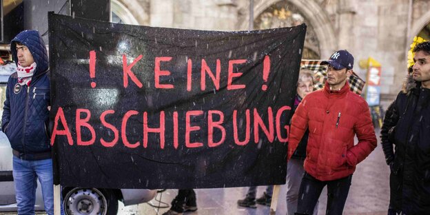 Drei Männer stehen im Freien neben einem Protestschild auf dem steht: Keine Abschiebung Drei Männer stehen im Freien neben einem Protestschild auf dem steht: Keine Abschiebung